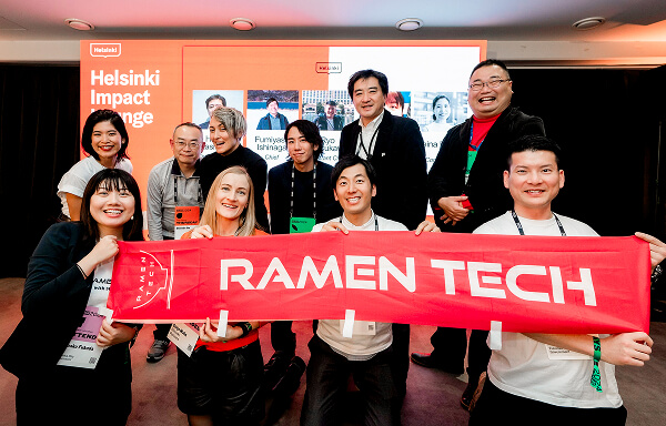 Helsinki Impactのイベントで、RAMEN TECHの赤いバナーを持った福岡のスタートアップ関係者たちが集合写真を撮っている。